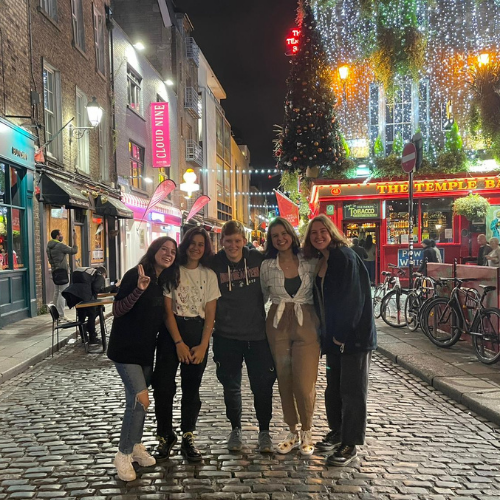 Five students standing in a cobbled street at night. Lights flicker in the background.
